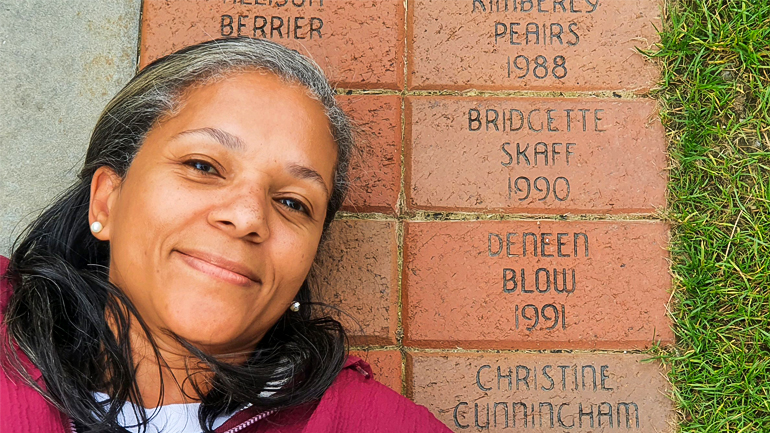 Deneen Blow is laying on the ground next to her named brick on the Quad. She is wearing a maroon jacket. 