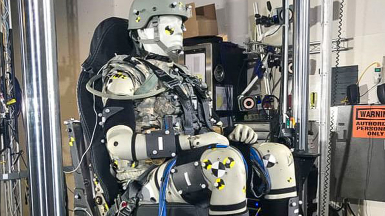 Pictured is a crash test dummy sitting in a seat and wearing a helmet. The Warrior Injury Assessment Manikin (WIAMan), the first vertical load assessment crash test dummy designed specifically to improve the safety of military vehicles by Henderson and a team at Diversified Technical Systems.