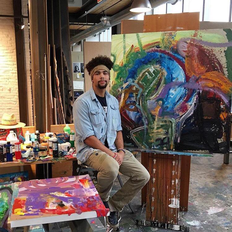 Merriman is in an art studio seated with a painting behind him